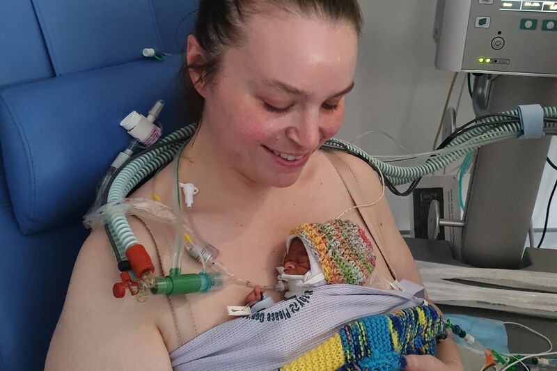 A parent in a hospital chair smiles looking down at their premature baby, who is directly against the skin on their chest, wrapped in a blanket. The baby has some medical tubes and wires attached to them.