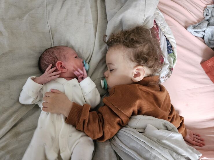 A young toddler and a newborn next to each other in bed, the older child has their eyes closed and their arm gently around the newborn.