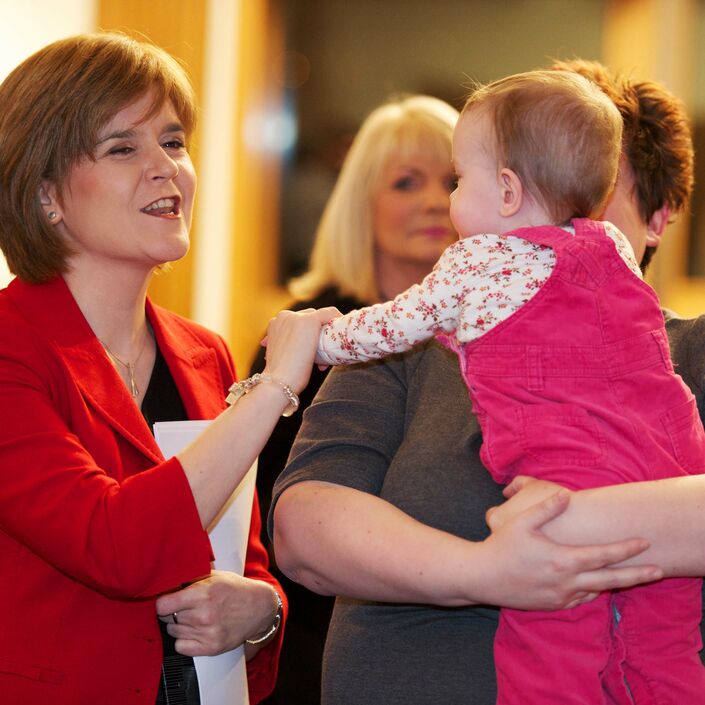 Mum holding toddler in her arms with Scotland's current First Minister (Nicola Sturgeon) holding the toddler's hand