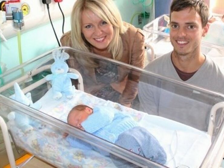 Dad and Esther Mcvey MP standing behind a cot with a premature baby smiling at the camera