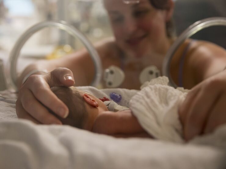 Baby in foreground in an incubator while mum in background has her hands through the incubator holding her baby's head