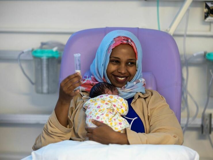 A mother sits smiling in a hospital chair, holding her small baby in her arms and a feeding tube in one hand