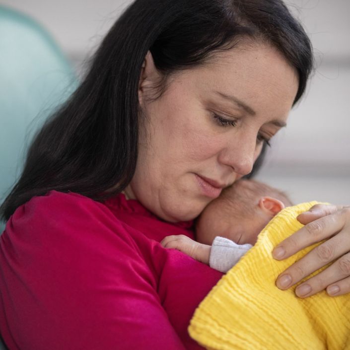 A mother sits in a hospital chair holding her small baby on her chest, who is wrapped in a yellow blanket.