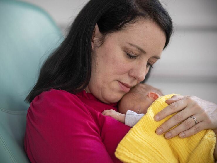 A mother sits in a hospital chair holding her small baby on her chest, who is wrapped in a yellow blanket.