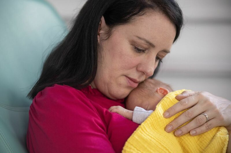A mother sits in a hospital chair holding her small baby on her chest, who is wrapped in a yellow blanket.