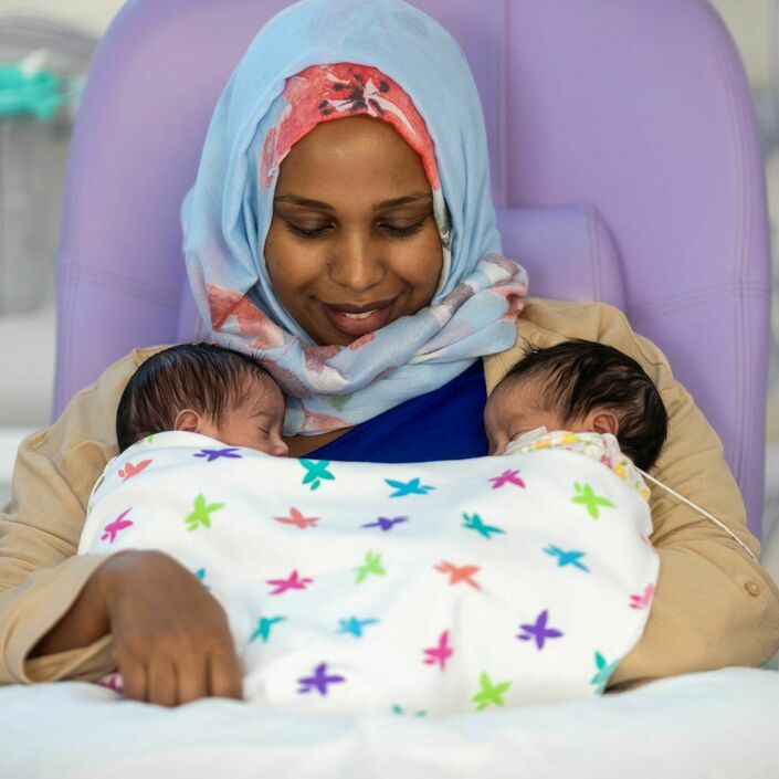 A mother sits in a hospital chair smiling down at her two babies in her arms, who are wrapped under a colourful patterned blanket. One baby has a wire attached to their face.