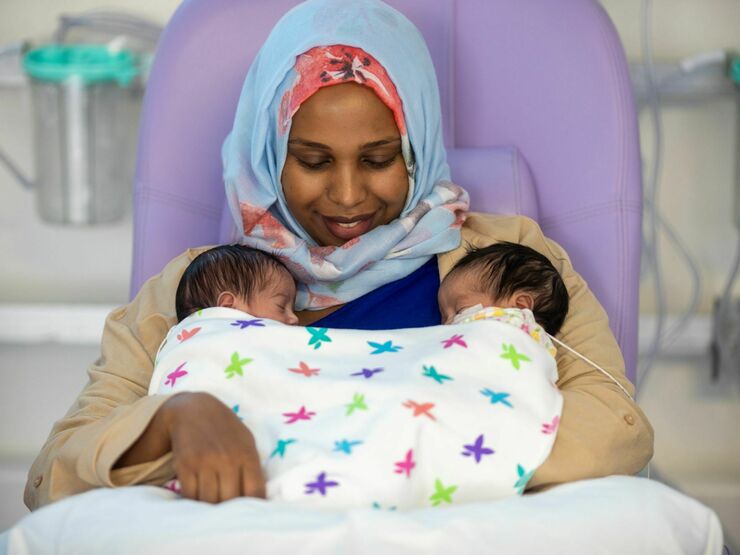 A mother sits in a hospital chair smiling down at her two babies in her arms, who are wrapped under a colourful patterned blanket. One baby has a wire attached to their face.