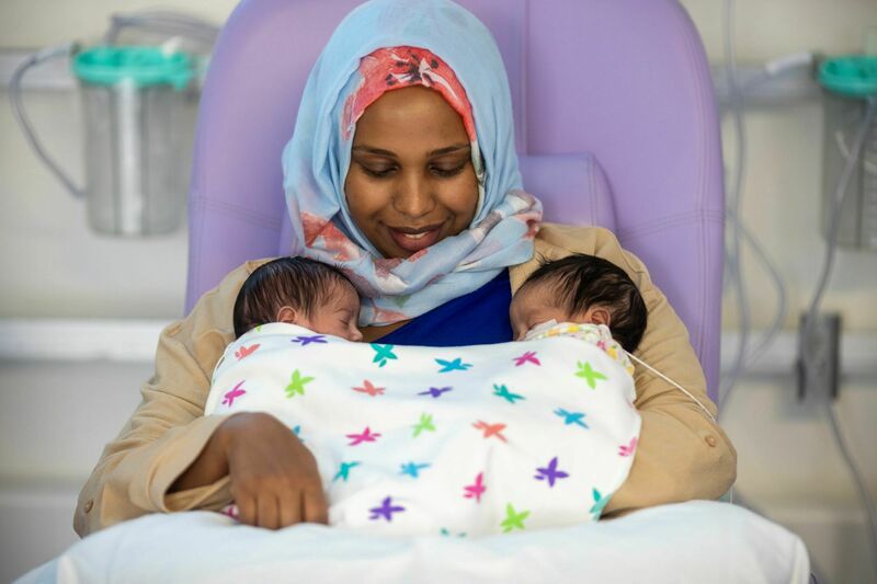 A mother sits in a hospital chair smiling down at her two babies in her arms, who are wrapped under a colourful patterned blanket. One baby has a wire attached to their face.