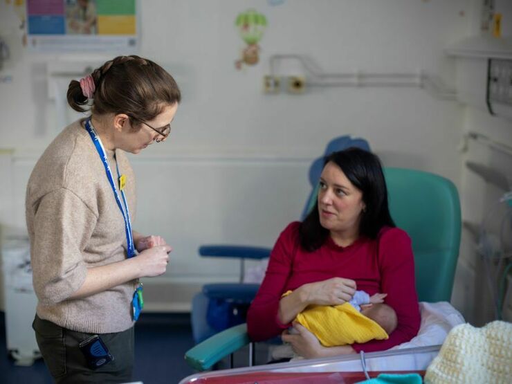 On a neonatal unit, a member of hospital staff and a parent are talking to each other, while the parent is sitting down holding a baby, wrapped in a yellow blanket.