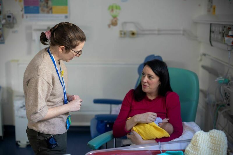 On a neonatal unit, a member of hospital staff and a parent are talking to each other, while the parent is sitting down holding a baby, wrapped in a yellow blanket.