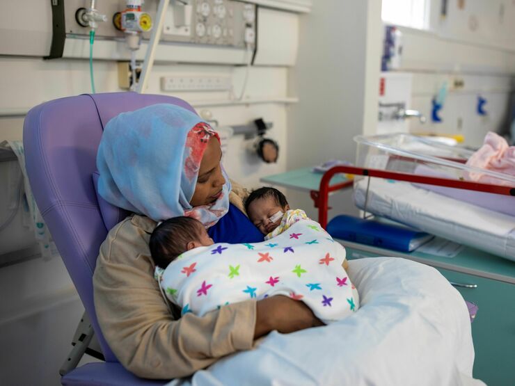 A mother sits in a hospital chair holding her two small babies on her chest, who are wrapped in a multicolour blanket.