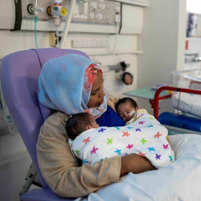 A mother sits in a hospital chair holding her two small babies on her chest, who are wrapped in a multicolour blanket.