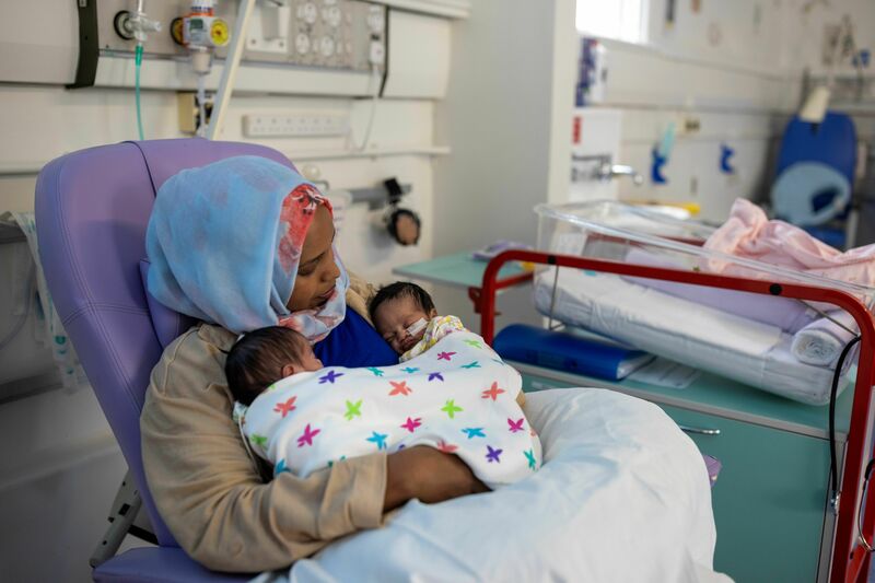 A mother sits in a hospital chair holding her two small babies on her chest, who are wrapped in a multicolour blanket.