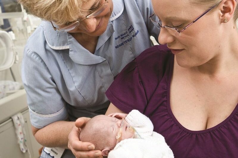 Mum holding baby in her arms with a nurse standing behind her with her hand supporting the baby's head