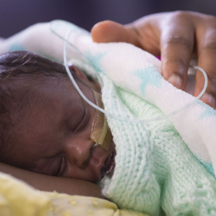 Premature baby wrapped in blanket sleeping on parent's chest with parent's hand on baby's back