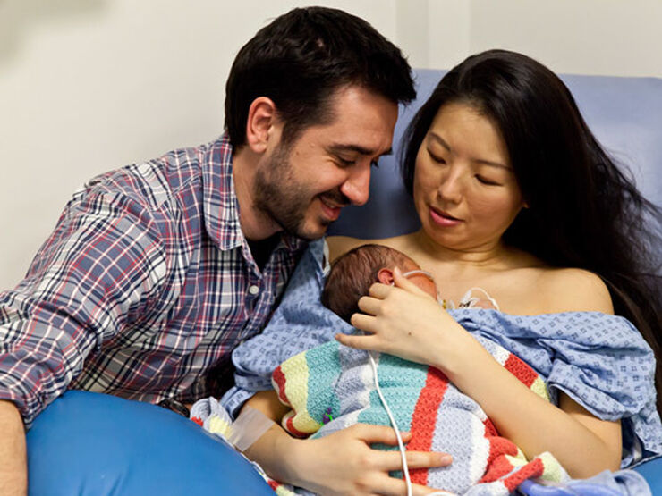 Mum holding baby on a hospital chair with dad alongside looking at baby. Baby is snuggled on mum's chest covered by a blanket.