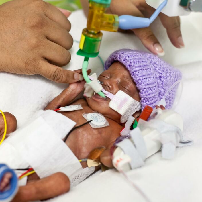 Premature baby in hospital wearing wool hat and attached to tubes holding parent's finger