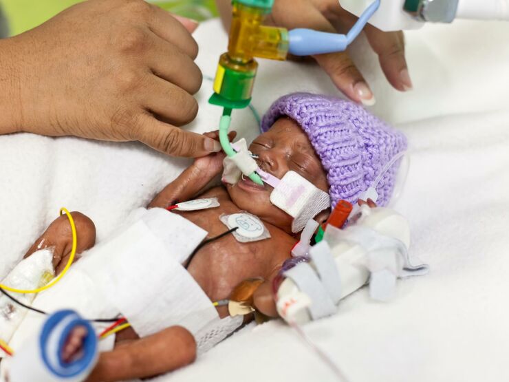 Premature baby in hospital wearing wool hat and attached to tubes holding parent's finger