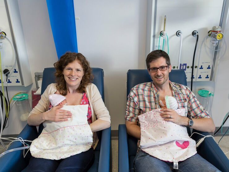 Mum and dad sat in hospital chairs smiling while holding twin babies