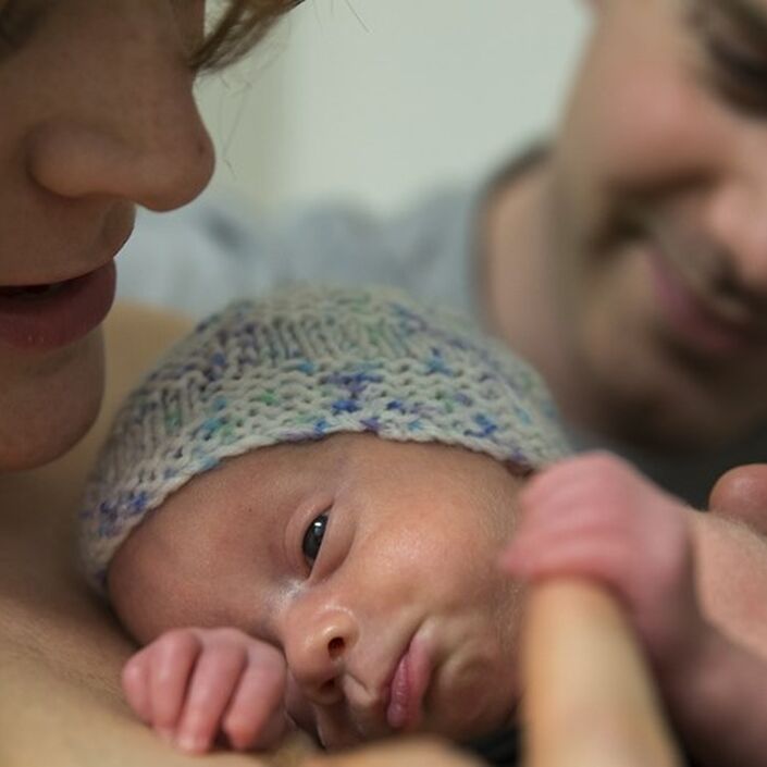 Baby skin to skin on mum's chest holding mum's finger and with dad in background with his hand on baby