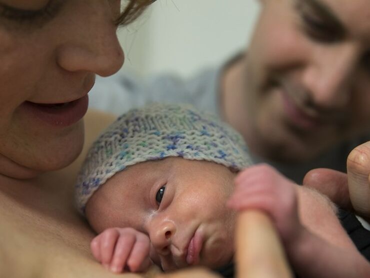 Baby skin to skin on mum's chest holding mum's finger and with dad in background with his hand on baby