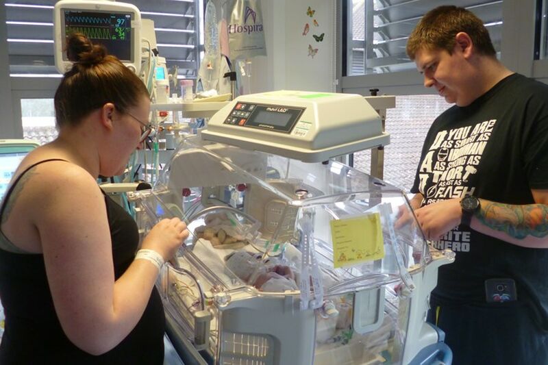 Mum and dad standing at either side of an incubator looking down at their newborn baby