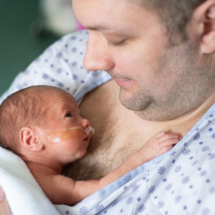 Dad in a hospital gown with his baby on his bare chest, the baby is looking up at him