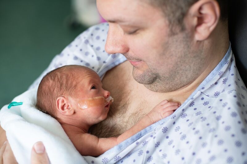 Dad in a hospital gown with his baby on his bare chest, the baby is looking up at him