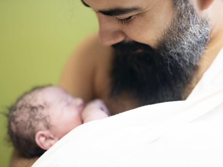 A bearded dad looks down and smiles at his baby who is on his bare chest. There is a blanket wrapped around one of his shoulders.