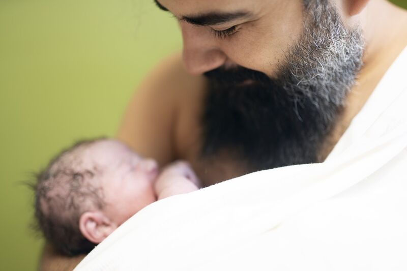 A bearded dad looks down and smiles at his baby who is on his bare chest. There is a blanket wrapped around one of his shoulders.