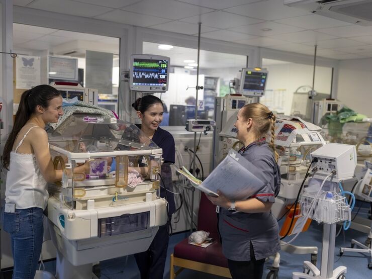 A parent in hospital standing next to her baby who is in an incubator, speaking to two members of neonatal staff. One of the staff members is holding a folder of paperwork.