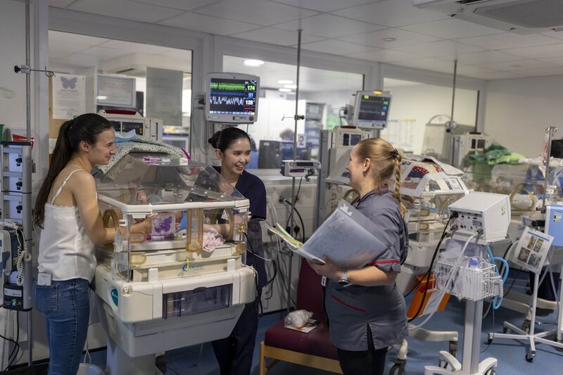 A parent in hospital standing next to her baby who is in an incubator, speaking to two members of neonatal staff. One of the staff members is holding a folder of paperwork.