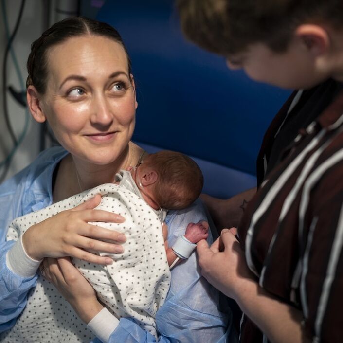 Parent in hospital chair holding a newborn baby, smiling up at another parent whose finger is being held by the baby.