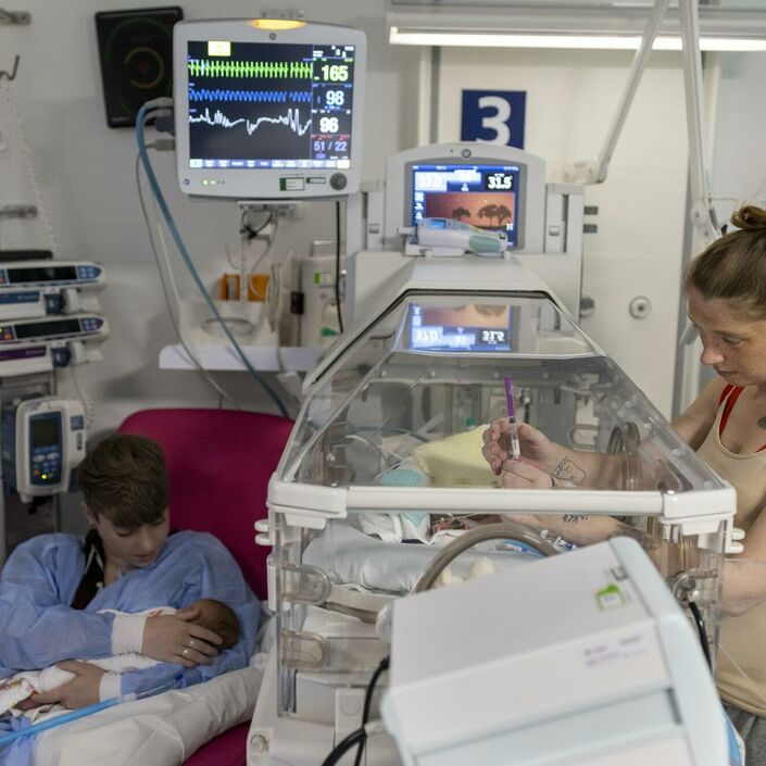 A parent is standing next to a baby in an incubator, with her hand through the hand hole touching the baby. Another parent is sitting in a chair next to the incubator, holding another baby.