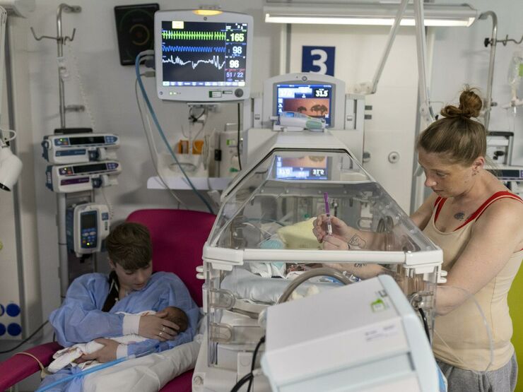A parent is standing next to a baby in an incubator, with her hand through the hand hole touching the baby. Another parent is sitting in a chair next to the incubator, holding another baby.
