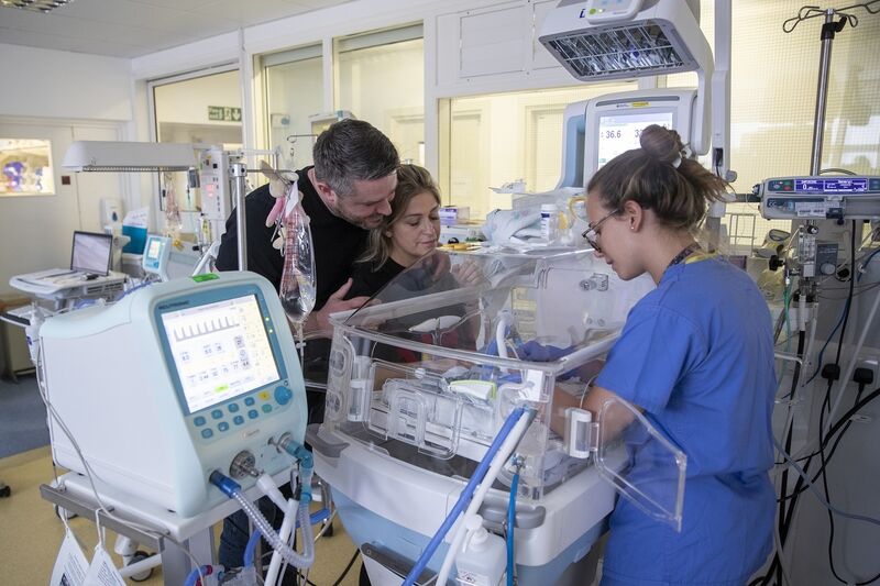 Parents with nurse in unit by cot looking at their baby while the nurse provides care for the baby