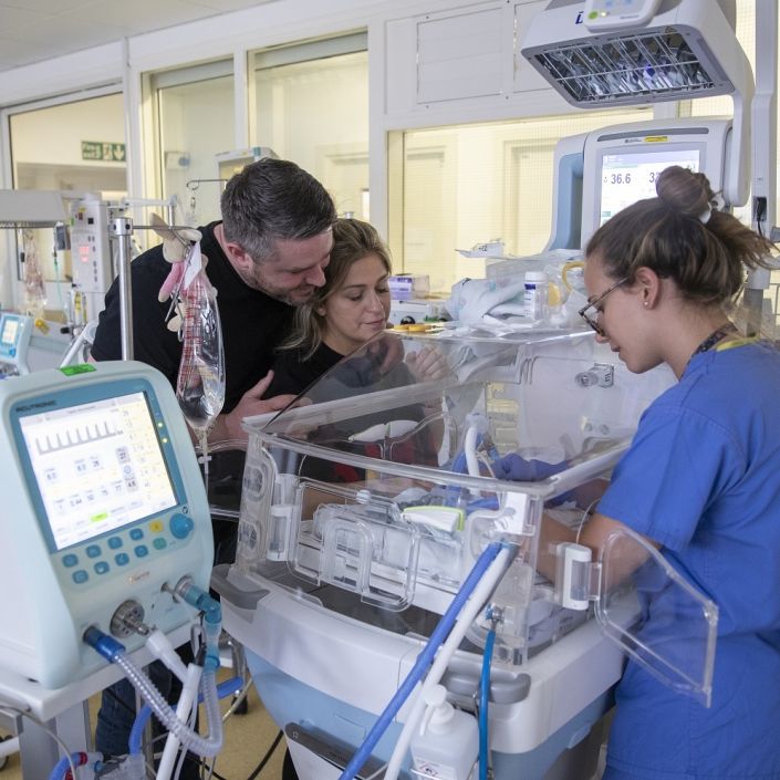 Parents with nurse in unit by cot looking at their baby while the nurse provides care for the baby