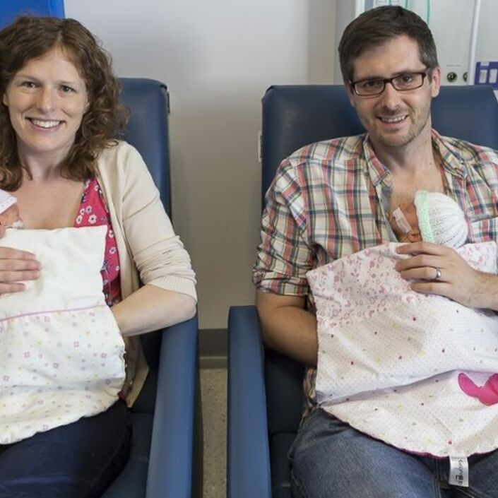 Mum and dad sitting in hospital chairs holding a baby twin each smiling at the camera