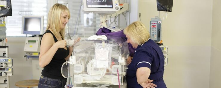 Mum and nurse either side of a cot looking and smiling at the baby