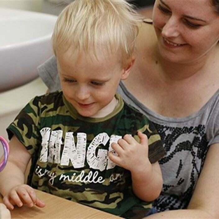 Little boy aged about 3 sitting on his mum at a table doing a task with health professional who has their back to camera