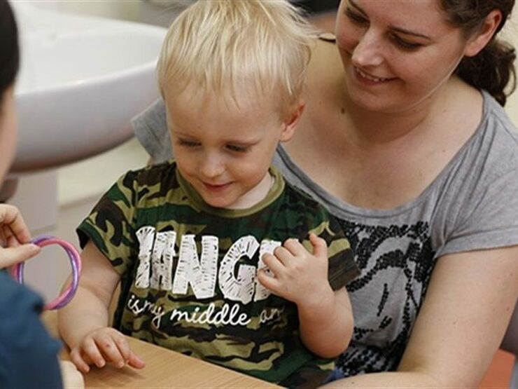 Little boy aged about 3 sitting on his mum at a table doing a task with health professional who has their back to camera