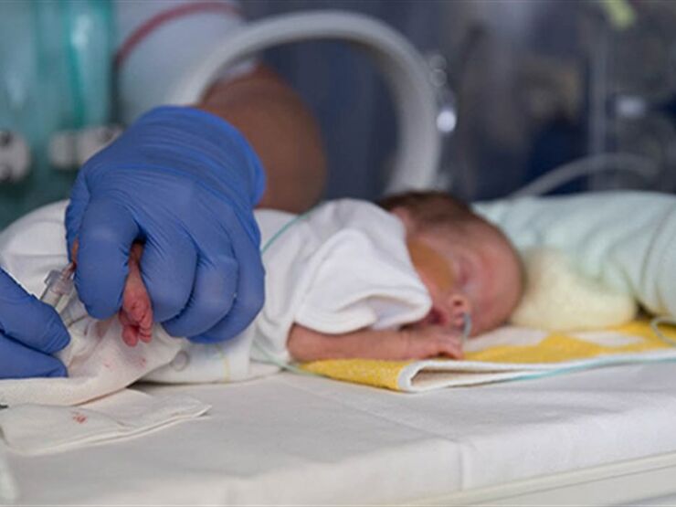 Close up of baby in cot lying on front with health professional wearing gloves touching the baby's feet