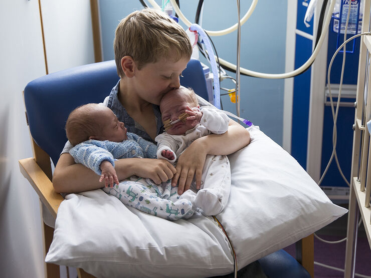 Twin babies being held by an older brother sitting on hospital chair with babies on a pillow and the older brother kissing one of their heads
