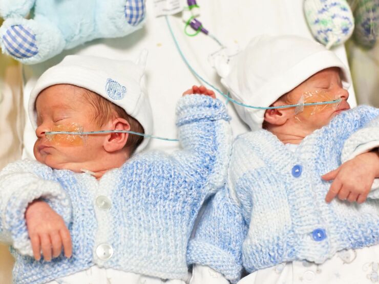 Twin babies asleep in hospital bed wearing blue cardigans and little hats with teddies above them