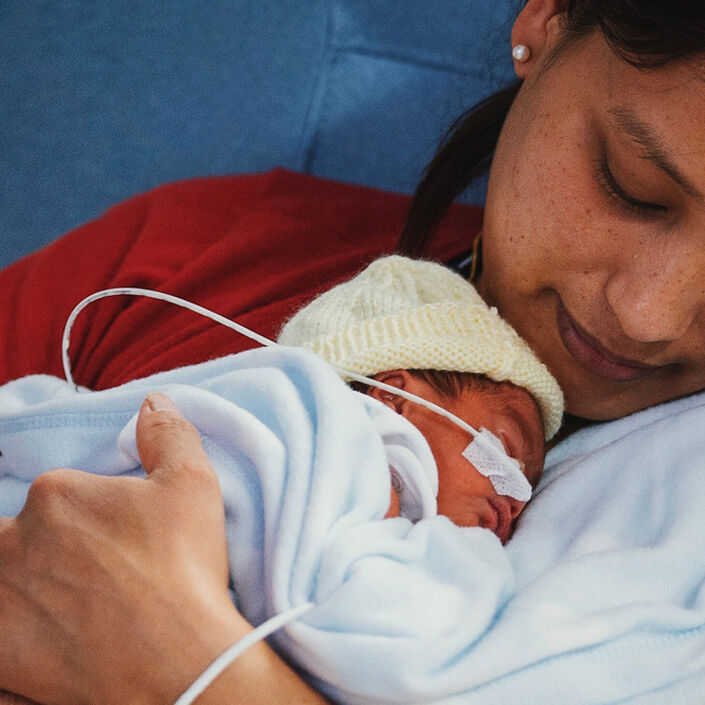Mother holding and looking down at her newborn on her chest. Baby wearing wool hat and being tube-fed