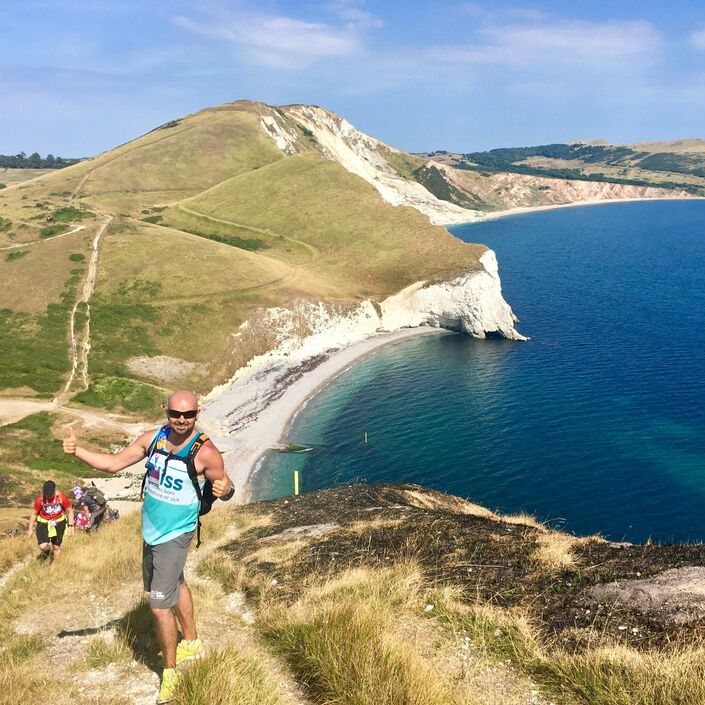 Man wearing Bliss top posing on the coast path with amazing sea views behind him
