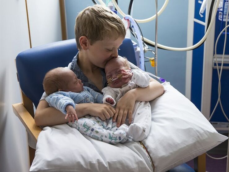 A boy holding his premature twin siblings in a hospital, kissing one on the head