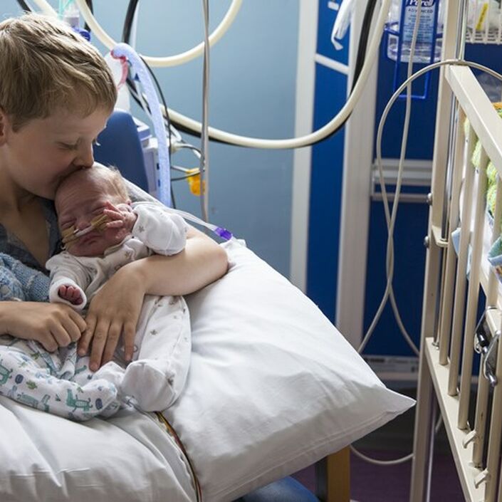 Brother holding twin siblings on his lap in hospital while kissing one on the head