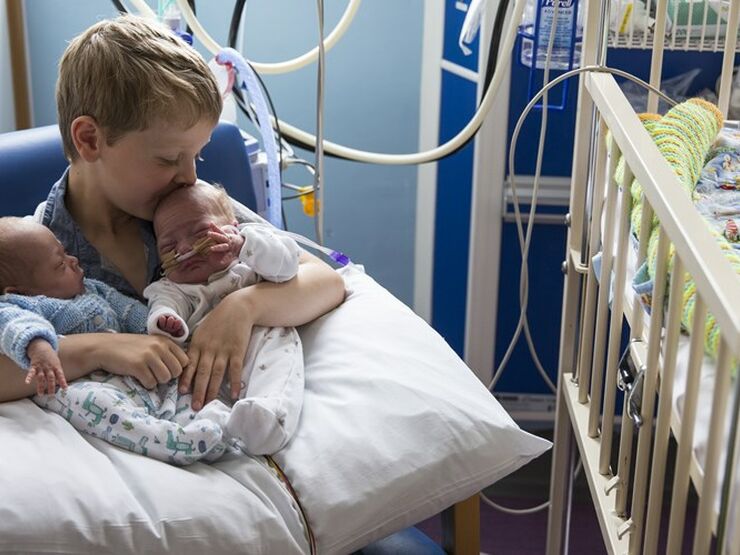 Brother holding twin siblings on his lap in hospital while kissing one on the head