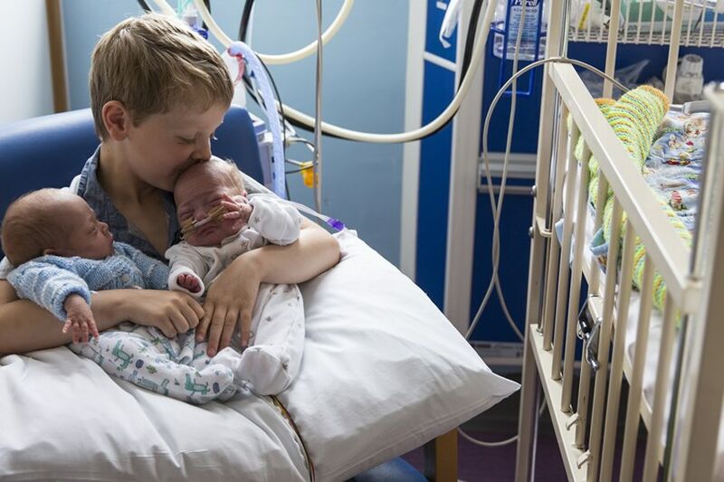 Brother holding twin siblings on his lap in hospital while kissing one on the head